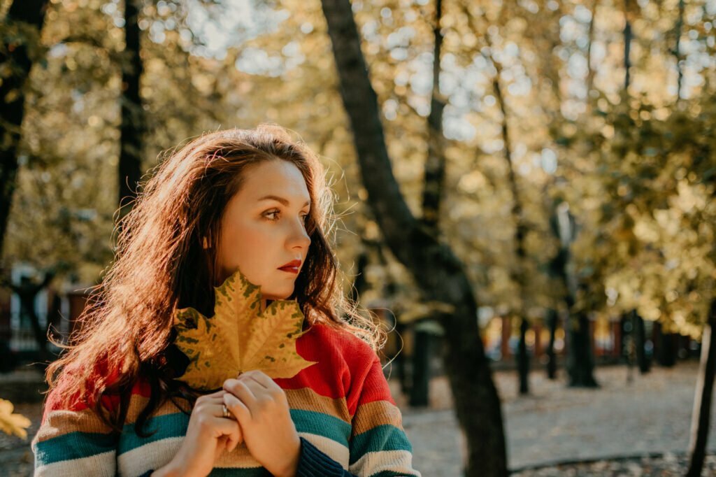 Woman holding a large yellow leaf, looking thoughtfully in a sunlit autumn park with vibrant