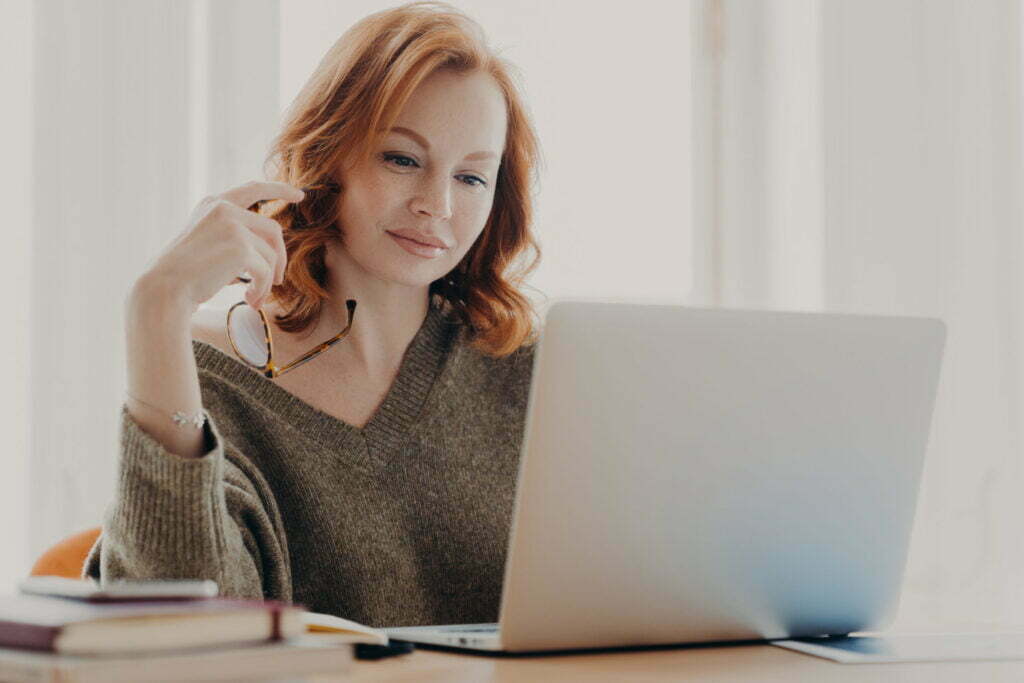 Woman with red hair in a cozy sweater using a laptop, focused on research and