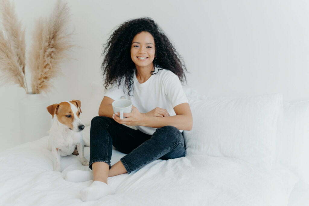 Smiling woman in casual attire holding a cup, sitting on a bed with a dog,